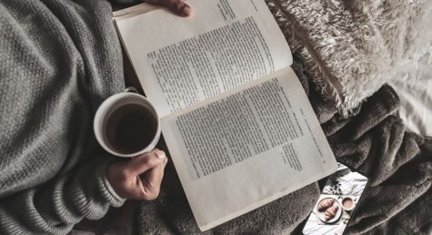 From above of crop anonymous female with mug of hot tea reading book under plaid near smartphone at home
